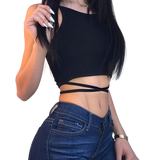 Close-up side view of a model wearing a black Femzai wrap-around crop top paired with blue denim jeans, accentuated by white nail polish and a gold bracelet, ideal for femboy clothing against a neutral wall background.