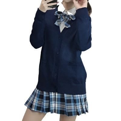 Medium shot of a model in a navy blue JK sweater paired with a blue plaid skirt and matching bow tie, representing Femzai's versatile femboy clothing line. The ensemble captures a youthful and preppy aesthetic against a simple indoor backdrop.