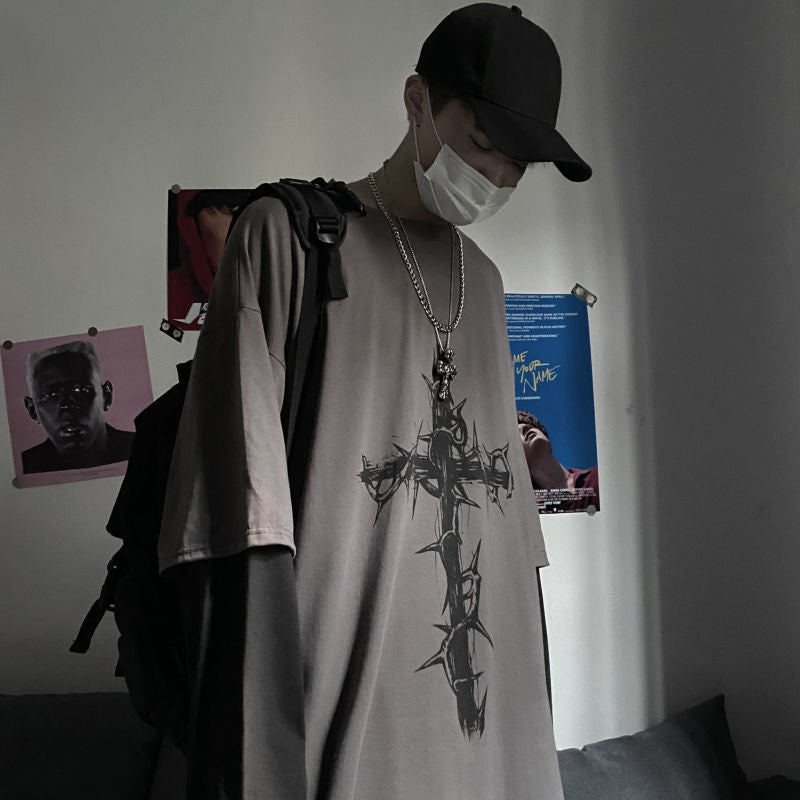 A medium shot from a higher angle showing a person in a femboy fashion outfit, featuring a long sleeve top with a cross double-sleeve detail. They are wearing a cap and face mask, with a chain necklace and pendant, complementing the edgy aesthetic.