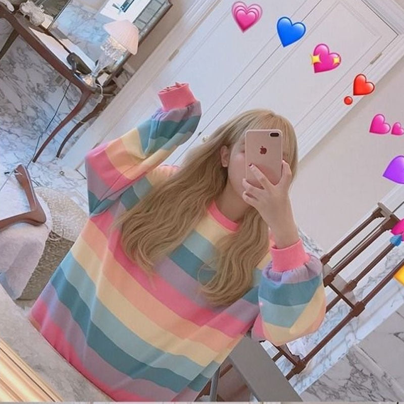 Indoor selfie shot with a high-angle view of a person in a mirror, wearing a femboy aesthetic rainbow-striped long sleeve shirt. The person's face is partially visible, holding a phone, with heart emojis floating around, adding a playful vibe to the image.