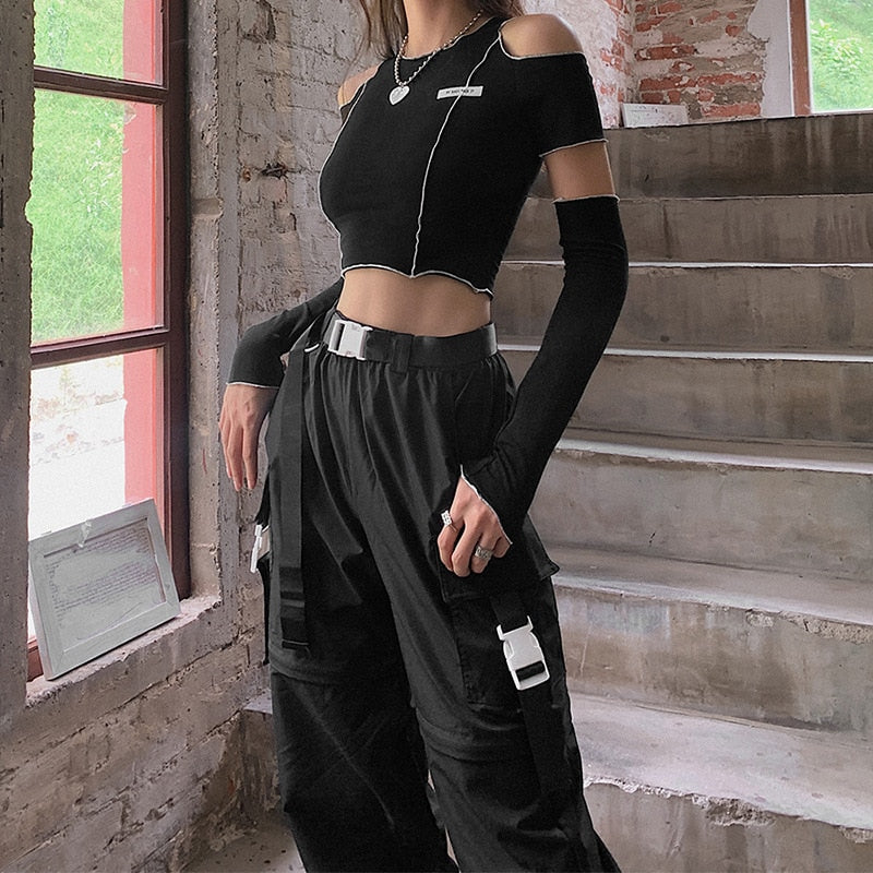 Product photo: Medium shot showcasing a femboy wearing the Femzai cut-out black crop top with long sleeves and high-waist pants, positioned beside a staircase with a rustic brick backdrop and a window revealing greenery.
