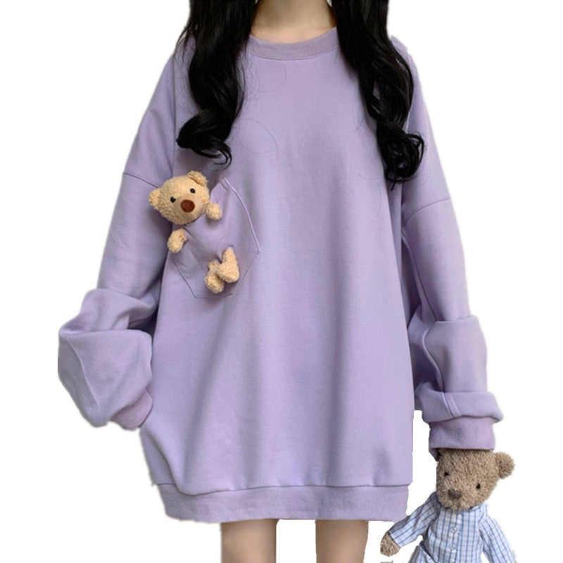 Image Description: Close-up shot of the Pouched Bear Sweatshirt in lavender. The design features a plush bear peeking out from a transparent chest pocket, with another bear hanging from the hem. The model's black wavy hair contrasts with the sweatshirt's pastel hue.