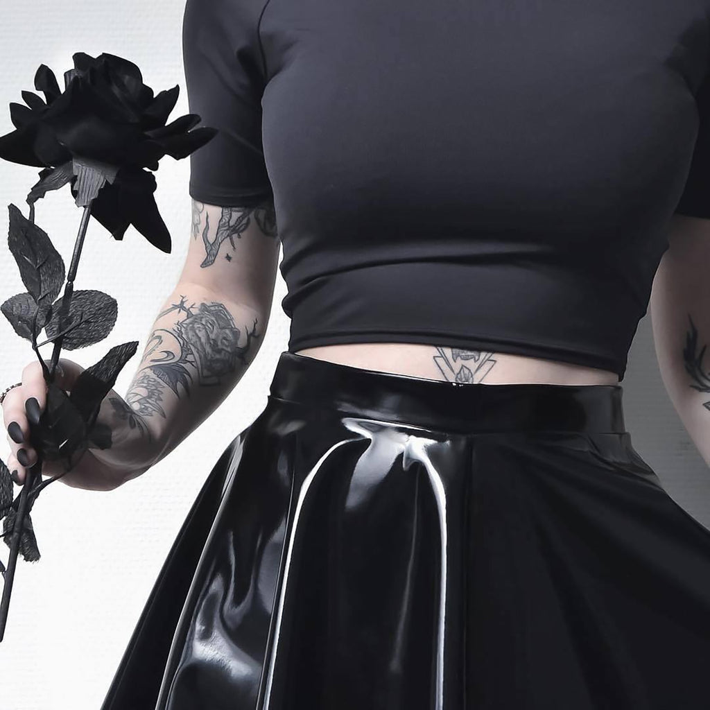 A femzai-inspired look showcasing a black form-fitting top and a glossy dancer mini skirt. The model holds a dark, artificial rose, adding a touch of gothic romance. Detailed tattoos on the forearm and wrist are prominently displayed, along with a minimalist tattoo on the lower abdomen. The entire aesthetic embraces an edgy, dark, and alternative style.