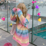 Outdoor shot of a person wearing a rainbow-striped long sleeve top in a femboy fashion style, standing near a glass door. The view is from a low angle, looking up as the person adjusts their hair with both hands, obscuring their face.