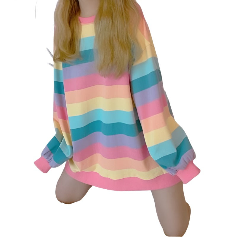 Image of a femzai model sitting on a white bed, wearing a femboy clothing style rainbow-striped long sleeve top. The shot is a high-angle view with a focus on the vibrant colors of the clothing and the person's crossed legs.