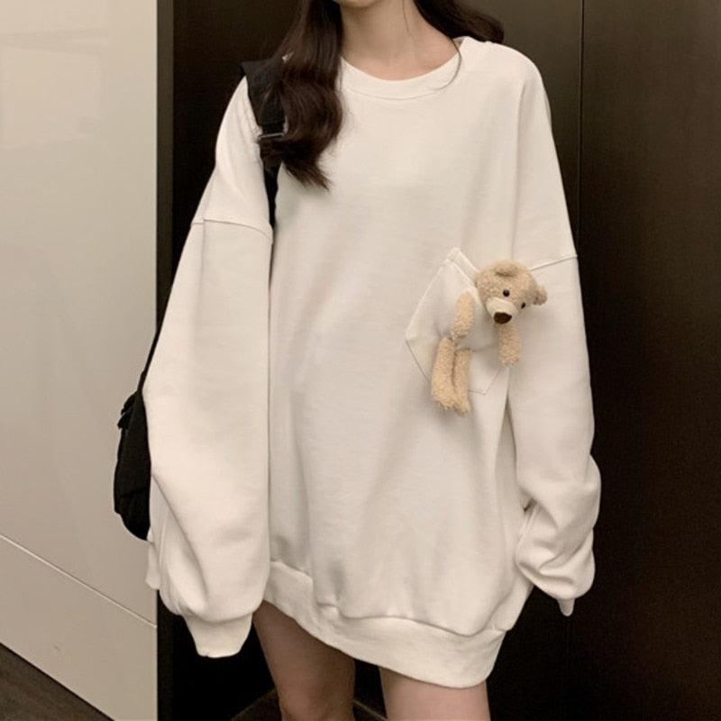 Image Description: Medium shot of the Pouched Bear Sweatshirt in white. The model, posed indoors, presents a side view accentuating the sweatshirt's relaxed fit. A plush bear is prominently visible from a transparent pocket, with the model carrying a black bag on her shoulder.