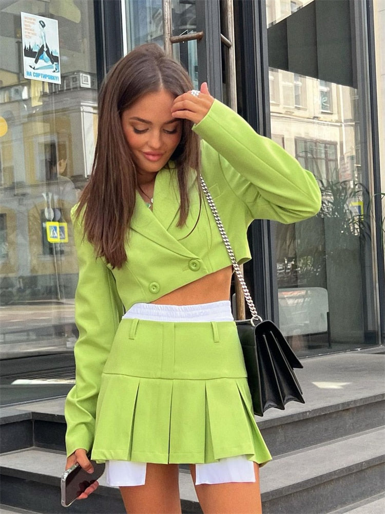 Medium shot view of a model in vibrant green femboy attire, showcasing a matching blazer and pleated skirt, standing in an urban setting.