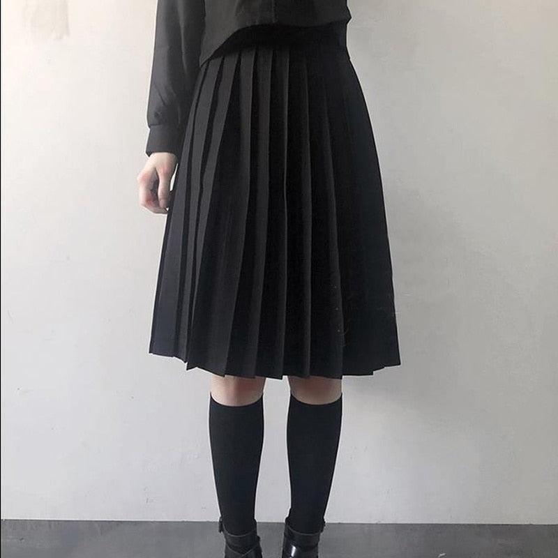 A person is standing with their hands by their side, wearing a long black pleated skirt, black knee-high socks, and black shoes. The image is a close-up shot focusing on the clothing, with a side angle view highlighting the femboy clothing style.