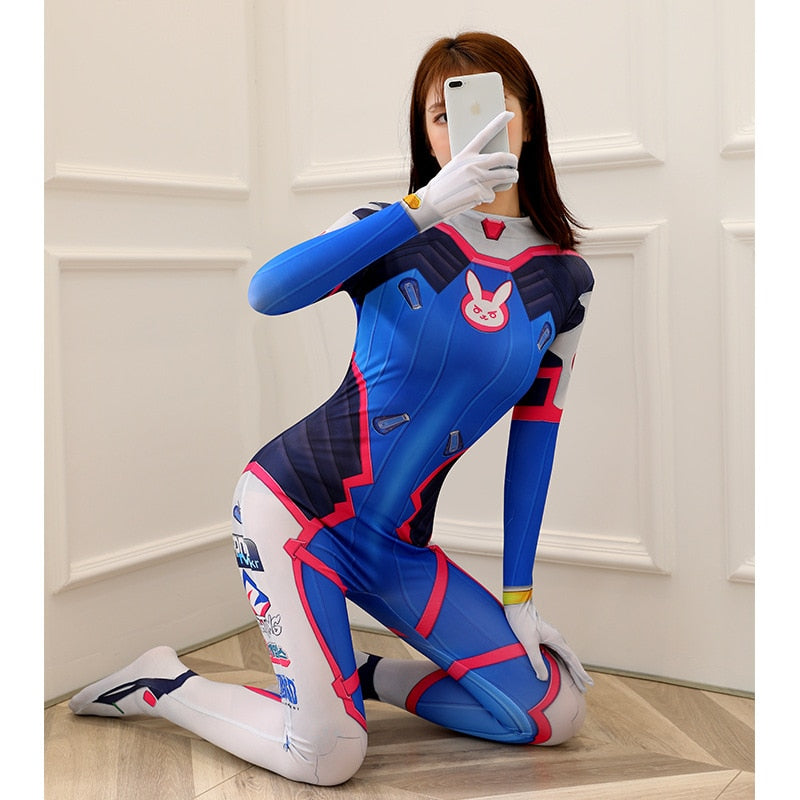 Medium shot front view of a model in a blue and pink Femzai D.Va cosplay suit, posing with a smartphone, highlighting the rabbit logo and other design details against a white architectural backdrop.