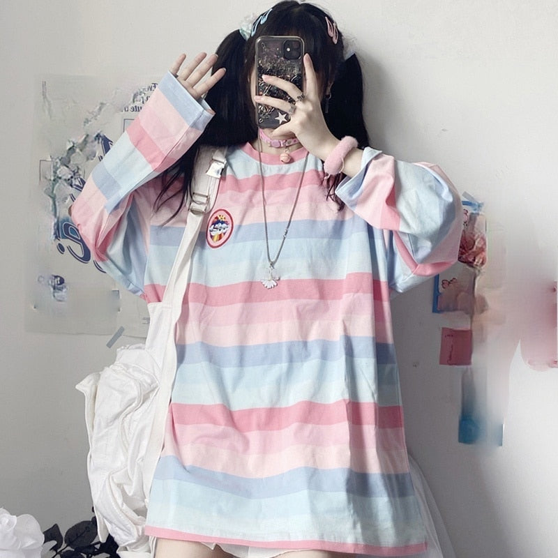 Medium shot with a front view of a model wearing a pastel-colored Femzai striped shirt, covering her face with a glittery phone case against a decorated wall, showcasing the shirt's relaxed fit and cute accessories.