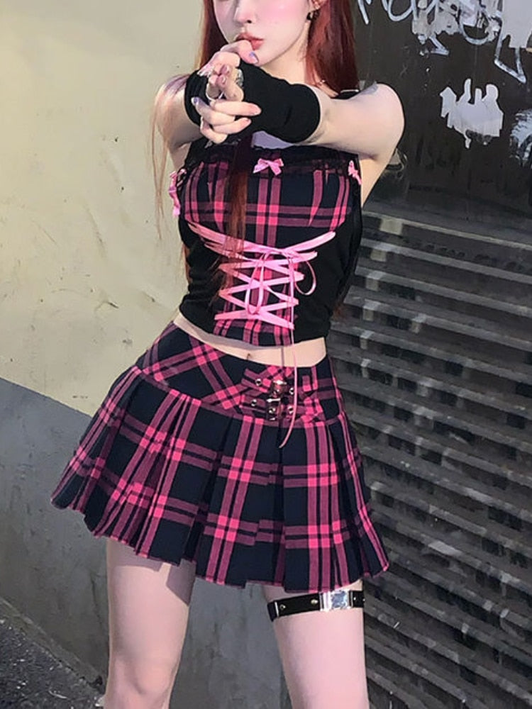 A femboy with vibrant red hair, striking a pose in a black and pink plaid off-shoulder top with intricate lace-up details. They're paired with a coordinating plaid mini skirt. The individual is adorned with bold black rings and confidently points towards the camera.