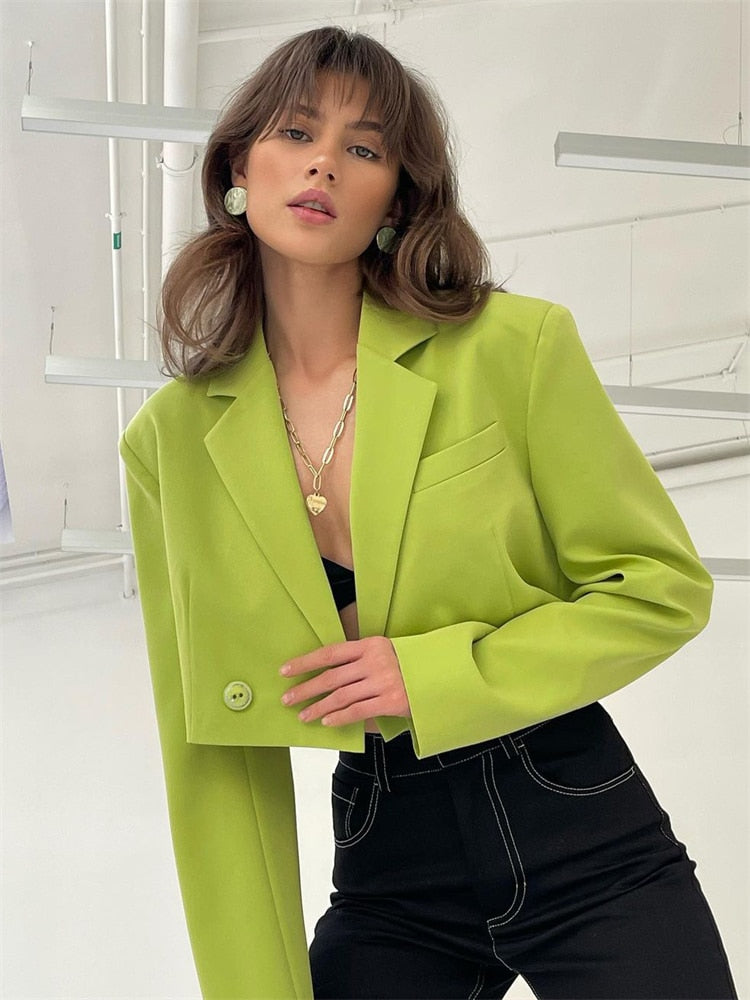 Medium shot view of a model in vibrant green femboy attire, showcasing a matching blazer and pleated skirt, standing in an urban setting.