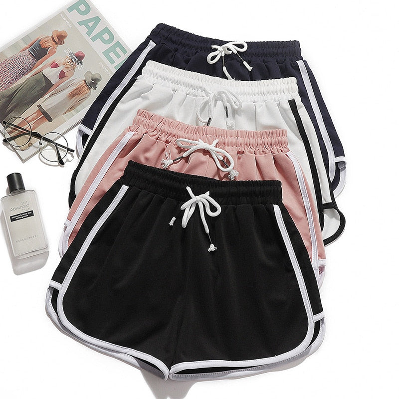 Flat lay view of a collection of femboy dolphin shorts in multiple colors: black, white, pink, and navy blue, paired with a magazine, glasses, and a bottle of lotion. Each short features contrasting side stripes and adjustable drawstrings.