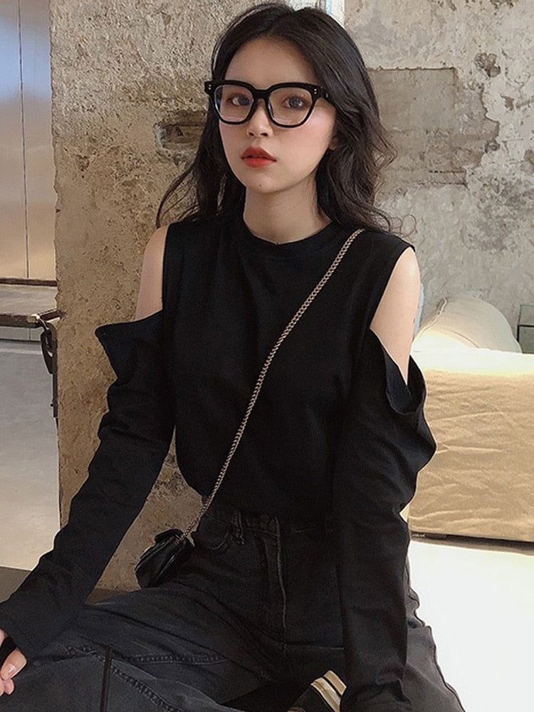 Medium shot front view of a model wearing black Femzai glasses, a black cut-out shoulder top with chain accessory, paired with black high-waisted pants against an urban rustic backdrop, perfect for chic urban fashion.