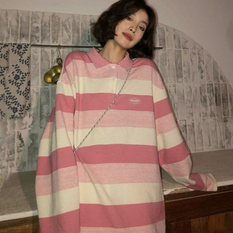 Side angled view of Striped Collared Longsleeve in pink and white. Model stands next to a distressed wall with blue floral designs, brass wall hook in view.