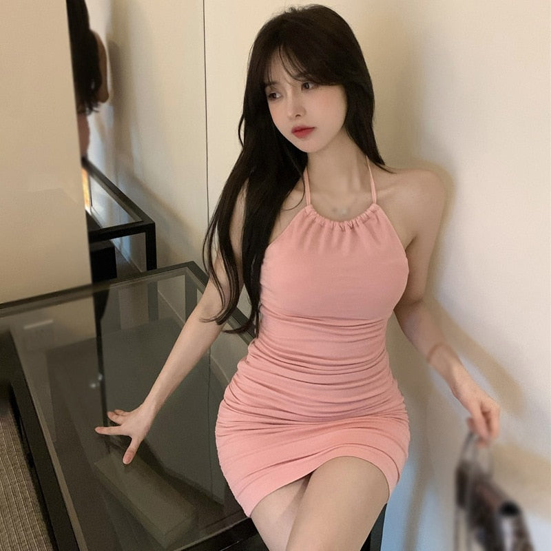 Image of a person posing in a room, wearing a pastel pink halter-neck mini dress, ideal for femboy clothing. The shot captures the individual from a front angle, providing a clear view of the outfit's fit and design.