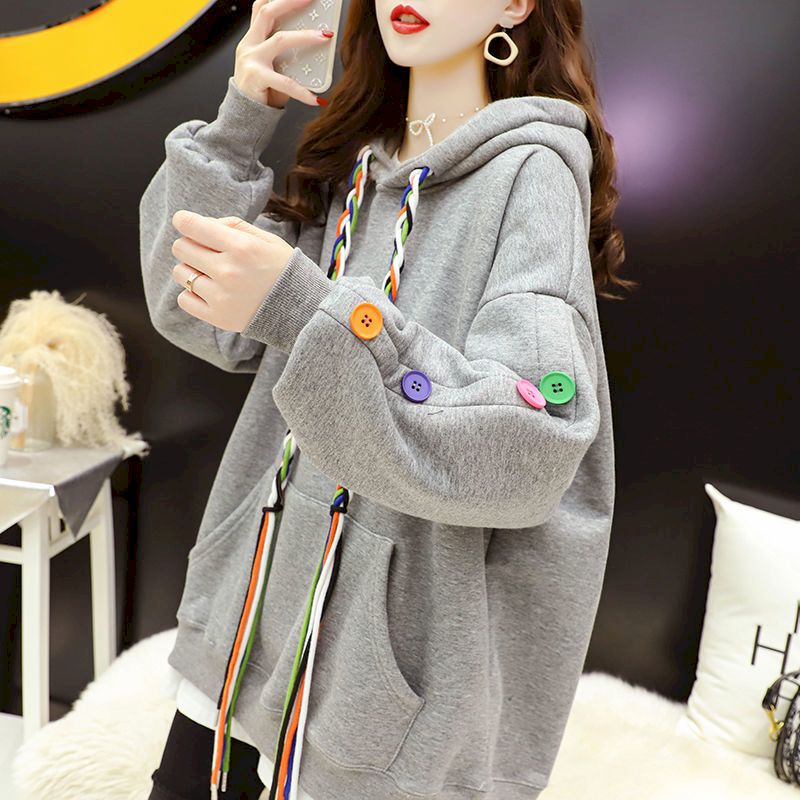 Medium shot of a model in a Femzai oversized gray hoodie with whimsical multicolored buttons and rainbow braided drawstrings, embodying a laid-back femboy clothing aesthetic. The model, holding a glass mug, is captured in a three-quarter view with a soft-focus background featuring chic interior decor, emphasizing the hoodie's cozy and fashionable appeal.