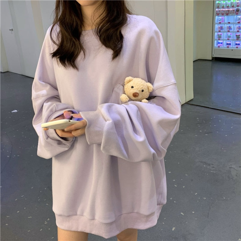 Image Description: Medium shot of the Pouched Bear Sweatshirt in lavender. A model stands in an indoor setting, holding a mobile phone, with a beverage vending machine in the background. The sweatshirt showcases a teddy bear peeking out from a transparent pocket.