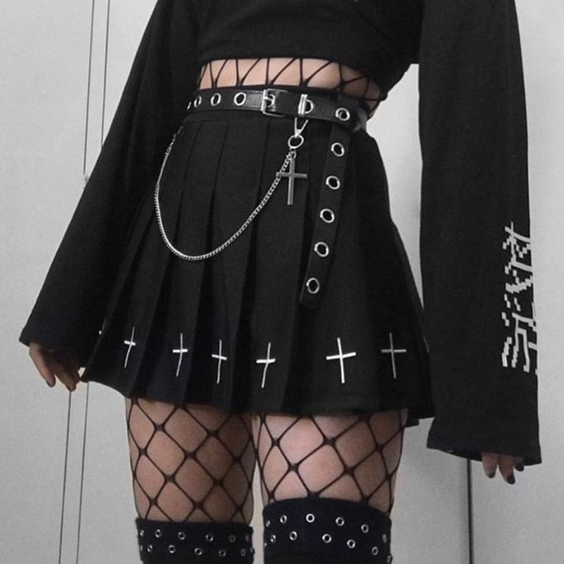 Detail shot of a femzai femboy clothing black alternative fashion ensemble; the outfit showcases a black pleated mini skirt adorned with metal rings, a silver chain with a cross pendant, and white embroidered crosses. Paired with the skirt, the model wears a black cropped top, black fishnet stockings, and studded garter belts. The sleeve of the top features white vertical typography, and the overall style exudes a punk-rock aesthetic.
