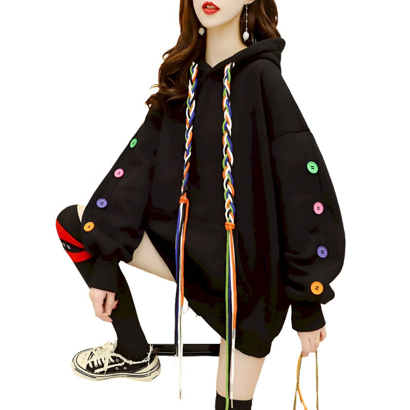Medium shot of a Femzai oversized black hoodie with colorful braided drawstrings and eclectic multicolored buttons, modeled in a side view that showcases a dynamic urban femboy clothing style. The model is striking a pose with a leg lifted, accentuating the relaxed fit of the hoodie against a modern indoor backdrop, with a hint of a vibrant orange chair adding a pop of color to the scene.